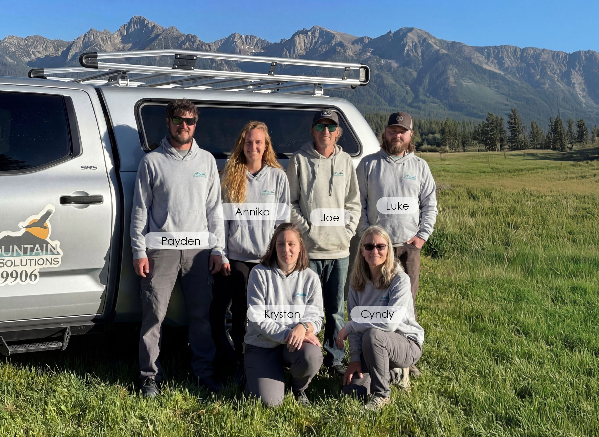 A team of six outdoor professionals poses in front of a silver truck with "Mountain Solutions" branding, against a backdrop of mountains.
